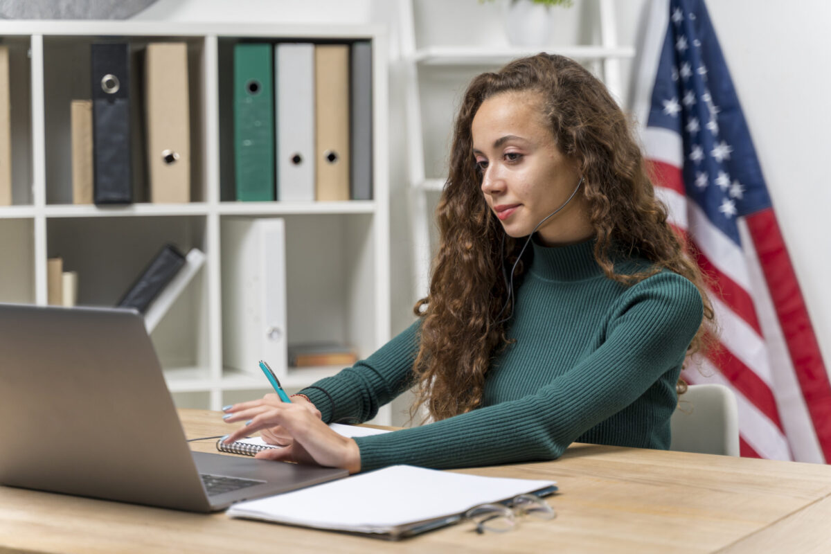 Fiatal nő tanul angolul laptopon keresztül, jegyzetel amerikai zászló előtt